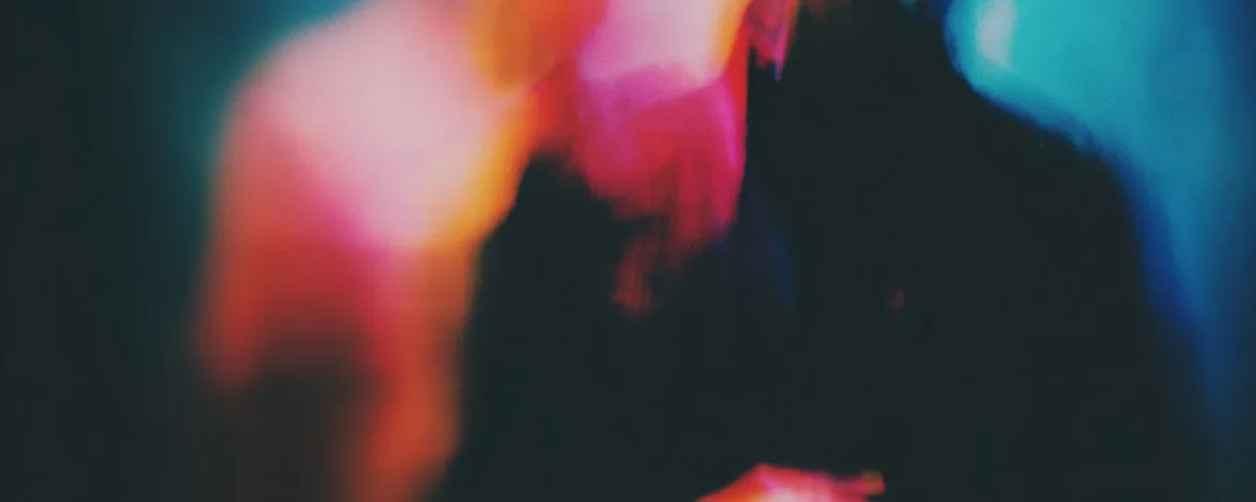 A soft-focus abstract in warm pink and cool teal light, a dark silhouetted form emerging from the blur.
