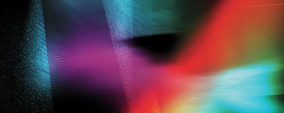 An abstract of smeared, motion-blurred prismatic light — teal, magenta, red, and orange streaks dragged across black around a dark central void.